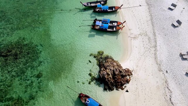 Explore the beauty of Koh Lipe, Thailand with a stunning aerial view of turquoise waters and traditional longtail boats gently bobbing in the waves. Relax on the sandy shores under the bright sun.