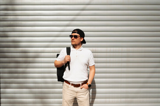 Man Posing Against Metal Shutter Backdrop, Neutral Expression, Sunglasses, Backpack Slung Over Shoulder,