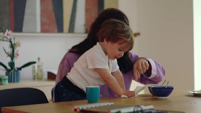 Toddler cleaning table with paper towel while mother guides by pointing showing learning moment and everyday parenting interaction in home kitchen environment
