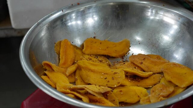 Market vendor mixes dried mango slices with red chili flakes by hand in a metal bowl, preparing a popular spicy fruit snack sold at Southeast Asian street markets. Street food.