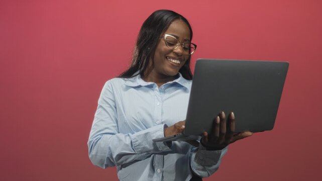 Woman holding laptop and typing with smiling face, wearing glasses and blue shirt in studio; confidence focus.