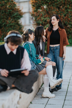 Three teenage students chat and laugh together outdoors on a college campus. A fourth student sits nearby using a tablet while the group socializes casually.