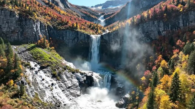 Stunning aerial view of a majestic waterfall cascading down a cliff surrounded by colorful autumn foliage and a rainbow