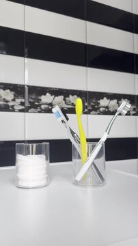 A hand picks up a yellow toothbrush and places it in a glass with several other toothbrushes in a bathroom