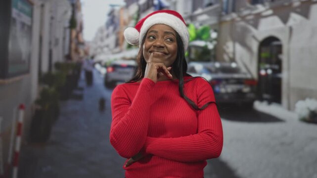 Woman wearing santa hat and red sweater with hand on chin and arms crossed on a snowy city street; festive joy.