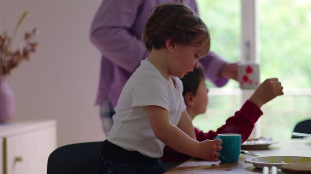 Mother serving drink to toddler at breakfast table while older child sits nearby creating authentic real life family moment in calm home environment