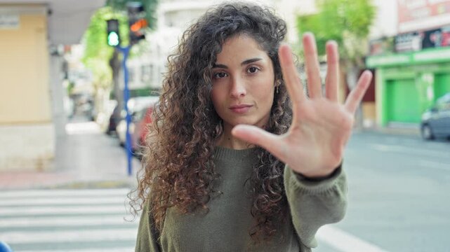 Woman with hand raised, palm forward stop gesture at a city street crosswalk wearing green sweater, curly hair framing face; defiance.