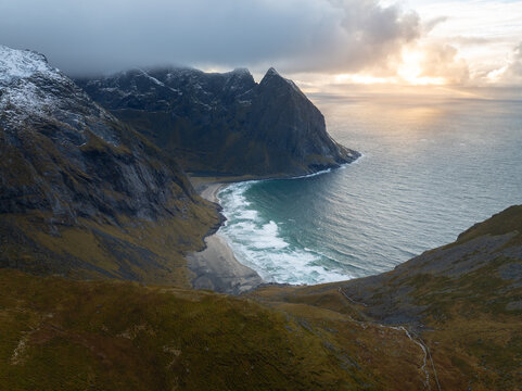 Aerial view of Lofoten islands featuring steep snow-dusted mountains overlooking a secluded sandy beach and the turquoise Norwegian Sea under dramatic clouds in Nordland, Norway.