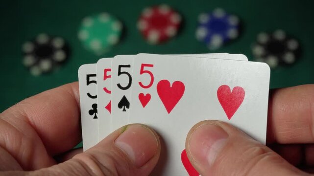 Close-up of hands holding playing cards showing four fives or quads, with poker chips in various colors blurred in the background on a green felt table