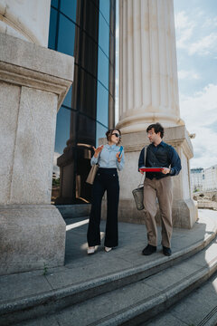 Two business coworkers walk and talk while reviewing documents and a tablet. The professional colleagues continue their discussion outside a large office building with classical columns.