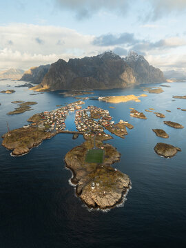 Aerial view of the Henningsvaer fishing village and its iconic football field on a rocky islet surrounded by the sea and rugged mountains in Henningsvaer, Nordland, Norway.