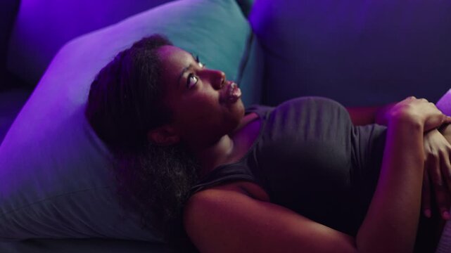 African American woman lying on couch staring at nails with bored expression passing time with idle gesture in quiet home interior during slow uneventful moment