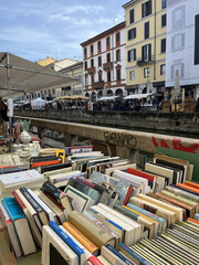 Fototapeta premium Photo of an outdoor antique and book market set along a canal in an urban area. In the foreground, rows of second‑hand books with colorful spines are displayed on tables under temporary stalls.