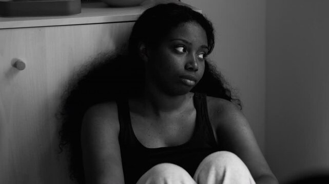 African American woman sitting on floor by cabinet looking sideways with tense expression showing depression anxiety emotional exhaustion feeling trapped, monochrome