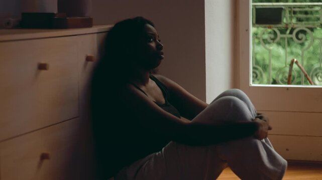 African American woman sitting on floor by window hugging knees with distant gaze expressing depression isolation hopelessness and emotional detachment in quiet home interior