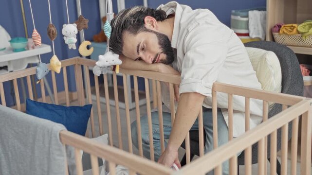 Man resting head on crib rail and reaching into wooden crib in a nursery room building; quiet fatigue.