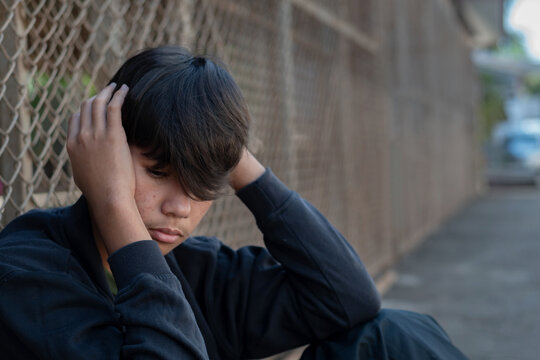 A teenage Asian boy sitting alone indoors, resting his arms on his knees with a sad and contemplative expression. Soft natural light and shallow depth of field emphasize his face and realistic skin te