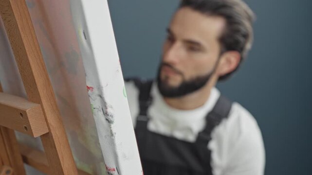Man painter in apron looks intently at canvas on an easel in studio, head tilted studying paint marks while wearing white shirt and black apron; concentration.