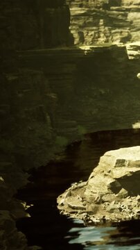 A serene river winds through towering stone cliffs, reflecting golden sunlight. Lush greenery peeks from the rocky terrain, creating a tranquil scene of natures beauty at dawn.