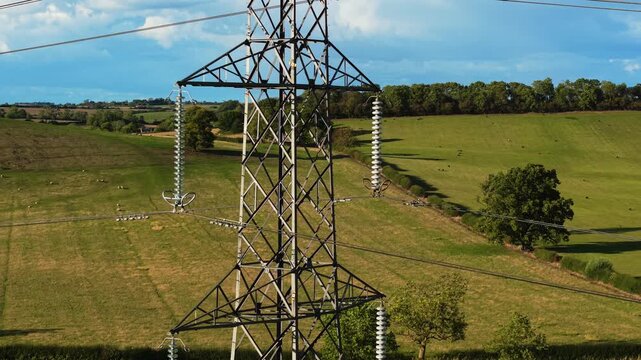 High voltage pylon rising above rural meadow. Large electric transmission poles transmitting electricity. Tall lattice tower carrying overhead current conductors over countryside field