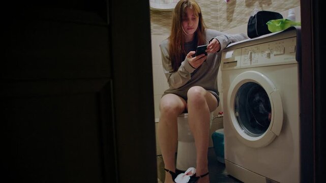 Witness shot of a young woman with period using her smartphone while sitting on the toilet in the bathroom at home
