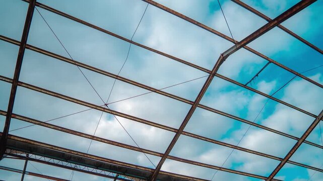 Abandoned hangar interior skeletal metal trusses under drifting clouds time lapse. Corroded framework decay atmosphere sky motion through broken roof timelapse. Urban exploration ruin neglect post