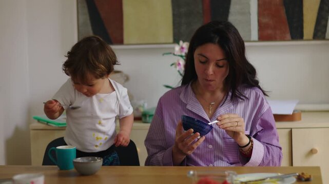 Mother and young child sharing breakfast at table, casual morning routine with playful mess, nurturing interaction, and everyday family life in warm domestic kitchen setting