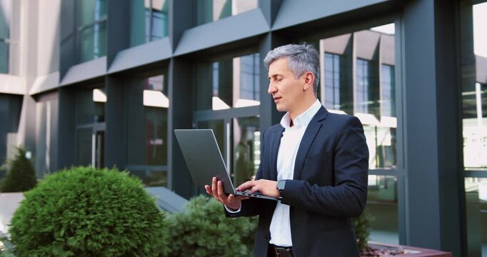 Handsome businessman working on a laptop outdoors near a modern glass office building, representing remote work, digital communication, and a modern urban business lifestyle
