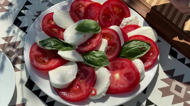 Caprese salad on white plate, sliced ripe tomatoes and mozzarella with fresh basil leaves, sunlit outdoor table setting with patterned cloth, fresh summer lunch