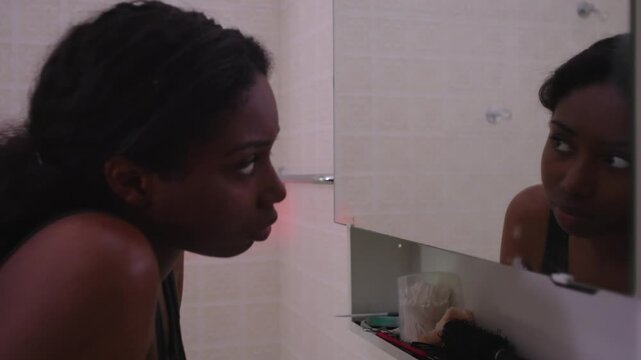African American woman examining face in mirror as camera pulls back revealing full body, touching skin with concern, expressing dissatisfaction, insecurity, and self criticism in bathroom