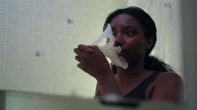 African American woman peeling off facial sheet mask in bathroom mirror, revealing fresh skin beneath, skincare routine moment focused on beauty treatment completion and self care ritual