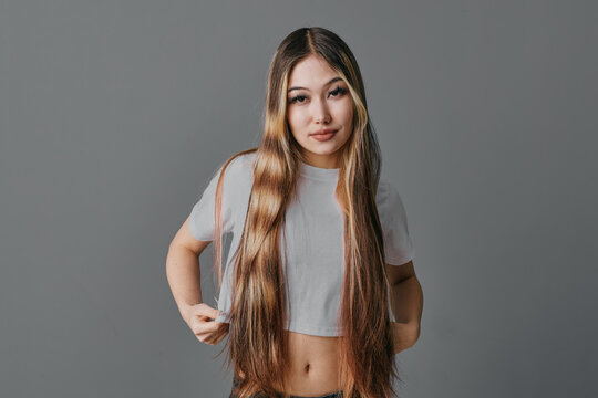 Young woman with long straight hair wearing cropped t shirt looks at camera with confident expression on gray background. Portrait of female beauty and casual style.