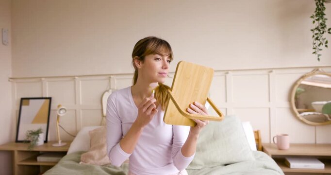 Woman in pajamas doing a facial massage with a jade roller while sitting on her bed in the morning