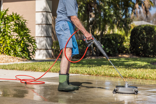 Man using pressure washer surface cleaner to power wash residential concrete driveway sidewalk.