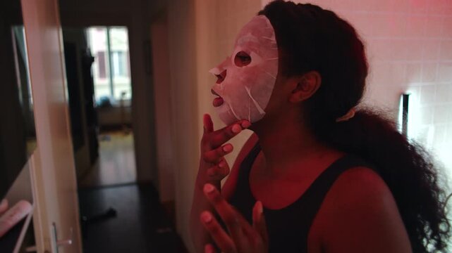 African American woman adjusting facial sheet mask on face while looking sideways, focused expression during skincare routine, quiet personal care moment in bathroom interior