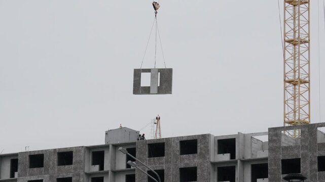 Hoisted wall panel lowered toward rooftop, worker signaling from roof edge, careful alignment with slot, tensioned cables, cloudy day