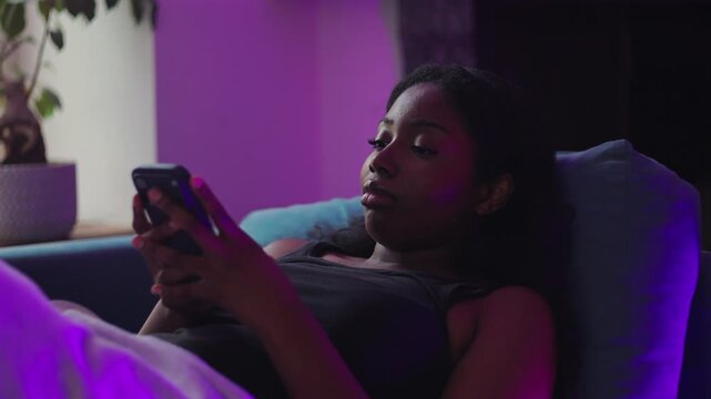 African American woman lying on couch sending voice message on smartphone, speaking with engaged expression, relaxed home environment and modern digital communication moment