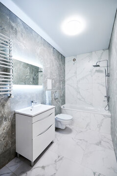 Contemporary bathroom interior with bathtub, suspended toilet and sink cabinet. Refined washroom with cool gray stone texture, white marble wall panels, backlit mirror and towel radiator