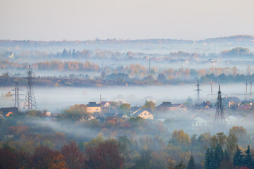 Obraz premium Fog drifts over suburban houses and autumn forest in layered morning landscape with power lines. Suitable for weather, environment, energy infrastructure, rural living or seasonal themes