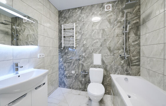 Bright bathroom interior with bathtub, sink vanity and toilet in gray stone finish. Contemporary washroom with rainfall shower, heated towel rail, lighted mirror and sleek design