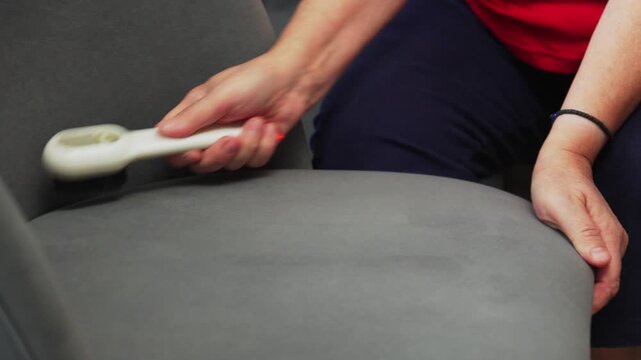 Person using a lint remover on a gray fabric chair to clean the surface while sitting on a couch in a living room