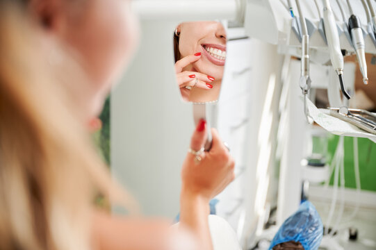 Female patient admires healthy teeth in mirror at dental office. Close up reflection with treatment unit in background. Useful for clinic advertising, cosmetic dentistry branding, wellness visuals