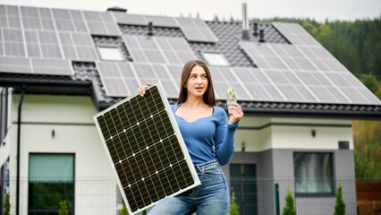 Woman holding solar panel and showing money near house with rooftop panels. Concept of renewable...