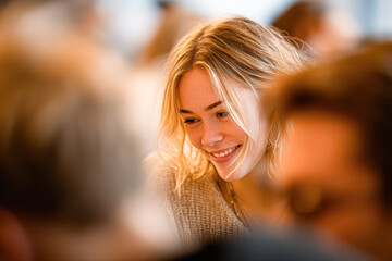 Obraz premium A young woman with blonde hair smiling warmly in a softly lit, social indoor setting.
