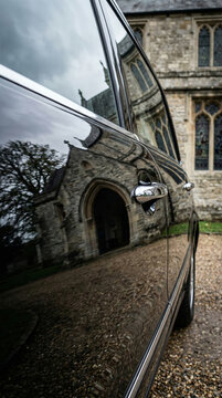 Black hearse parked outside stone church. Funeral ceremony for veteran and hero. Traditional burial service, mourning and sorrow. Reflection of old building on glossy vehicle exterior metal.