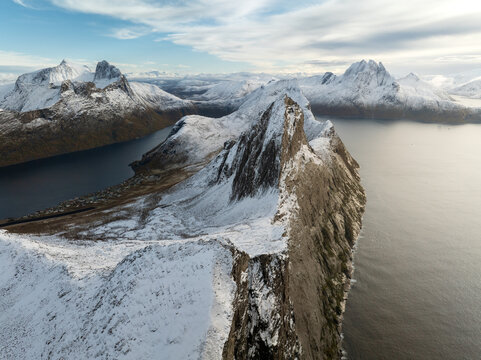 Aerial view of the iconic Segla mountain peak with its sharp snow-covered ridge overlooking the deep blue waters of the fjord and surrounding arctic peaks in Senja, Troms, Norway.