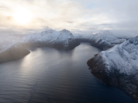 Aerial view of snow-covered mountains and the calm waters of the fjord in Husoy, Troms, Norway.