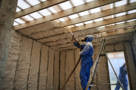 Installer covering overhead framing with spray foam insulation in house under construction. Ladder access, protective suit and energy saving building envelope concept