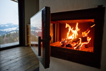 Naklejka premium Fire burns in modern fireplace inside cozy home. Close up of glowing logs and flames, glass door open, wooden floor and mountain view through window, warmth and comfort concept