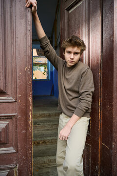 Teen leaning in doorway with gaze, intense eye contact, hand bracing doorframe, textured wood panels, warm interior behind, neutral sweater, confident model energy, cinematic color grading